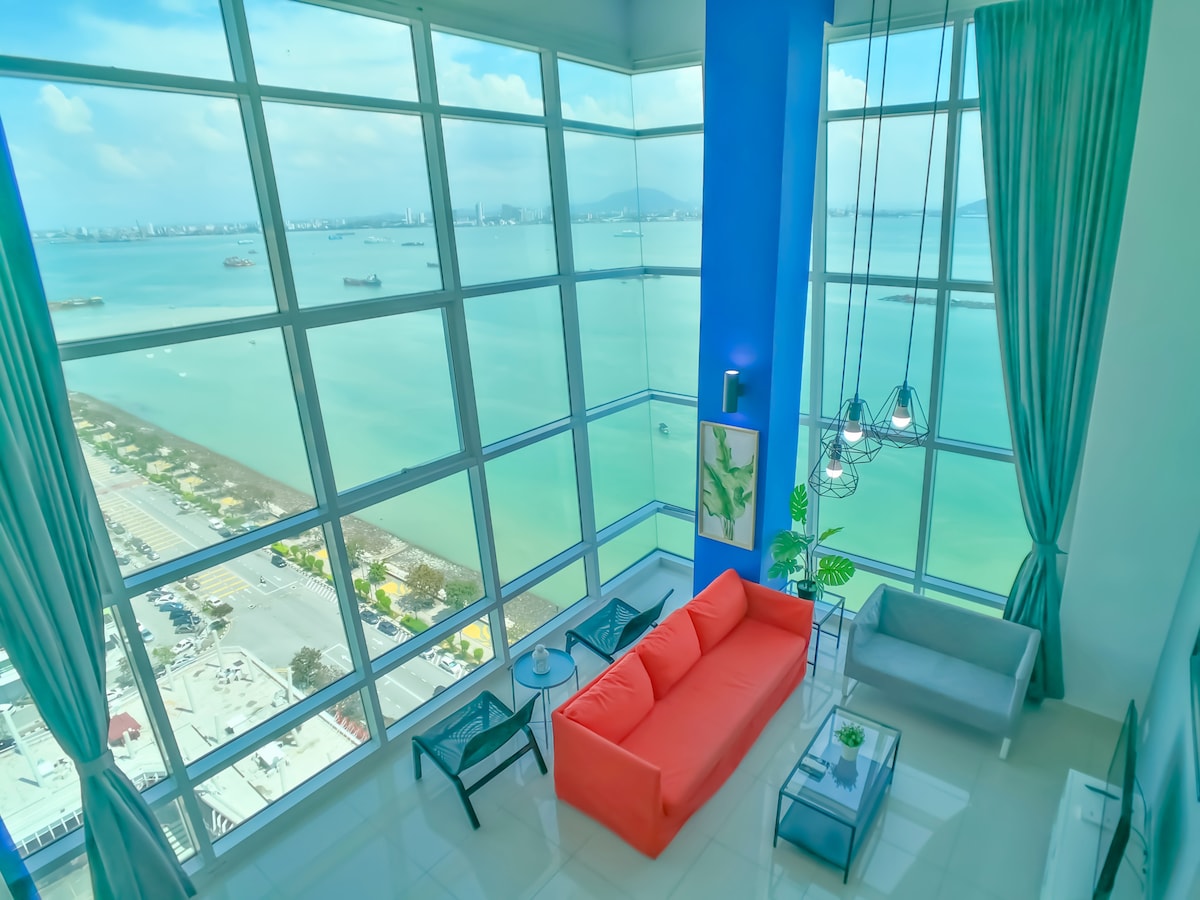 The spacious living area features large floor-to-ceiling windows, allowing for unobstructed views of the sea. A bright orange sofa is positioned beside modern glass tables, while turquoise curtains add a pop of color. Natural light floods the space, creating a refreshing ambiance.