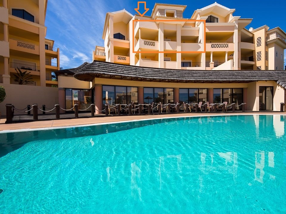A view of the resort shows a sparkling swimming pool in the foreground with clear blue water. Above, a spacious balcony is visible on the upper level of the building, framed by light-colored architecture. The outdoor seating area overlooks the pool.