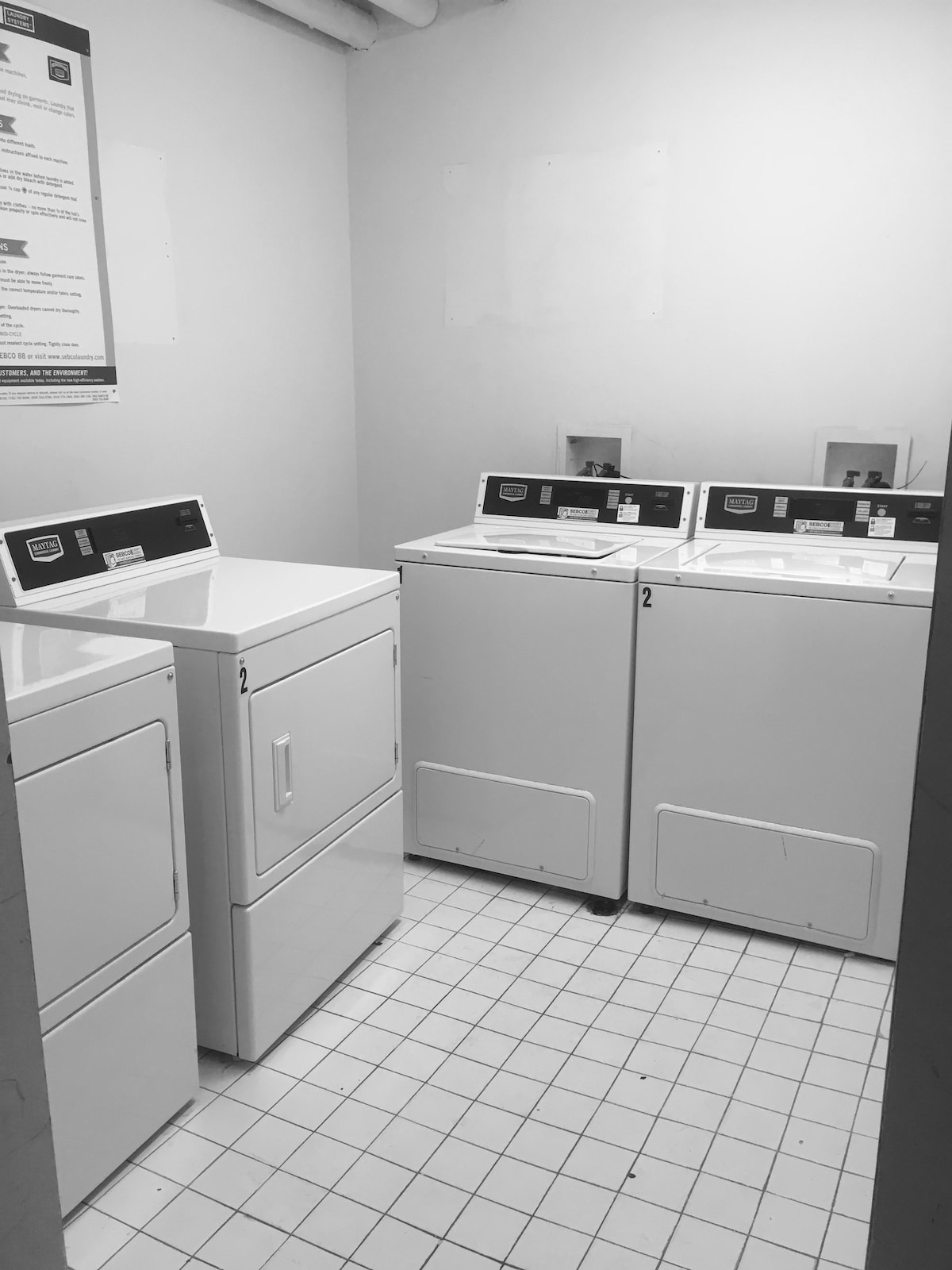 A clean laundry room features two washing machines and one dryer arranged against a tiled wall. Each machine displays a digital control panel. The space is well-lit, with light tiles covering the floor, contributing to a functional and practical area for laundry needs.
