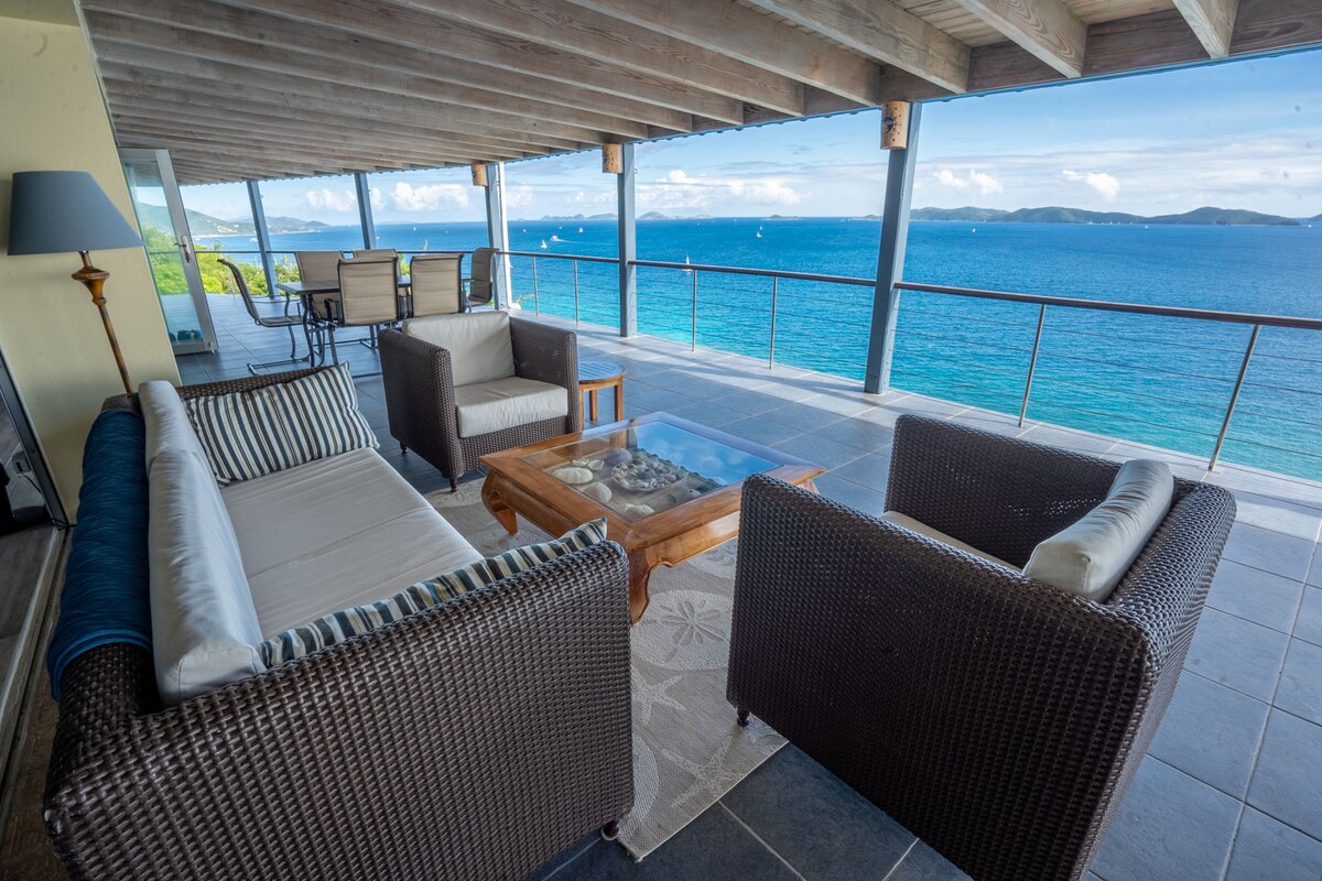 A covered outdoor deck features comfortable seating with wicker chairs and light cushions. A wooden coffee table sits at the center, with expansive views of the azure water and distant islands visible through the railings. Soft sunlight illuminates the open space.