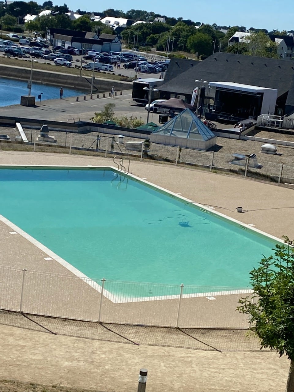 A clear swimming pool is visible, surrounded by a beige tiled area. The pool is enclosed by a white fence, and there are lounge chairs positioned nearby. In the background, an area with parked vehicles and a building can be seen, providing context to the surrounding amenities.