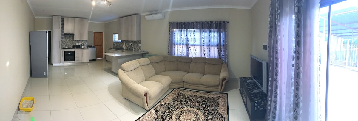 A spacious living area features a large sectional sofa arranged around a decorative area rug. Natural light fills the room through the window adorned with sheer drapes, while a modern kitchen can be seen in the background, showcasing cabinetry and appliances.