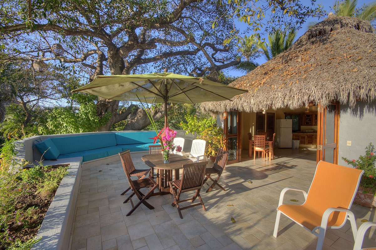 An outdoor deck features a thatched-roof palapa providing shade over a dining table surrounded by six wooden chairs. A large umbrella offers additional shade. Green plants and colorful flowers enhance the space, while a cozy seating area is visible in the background.