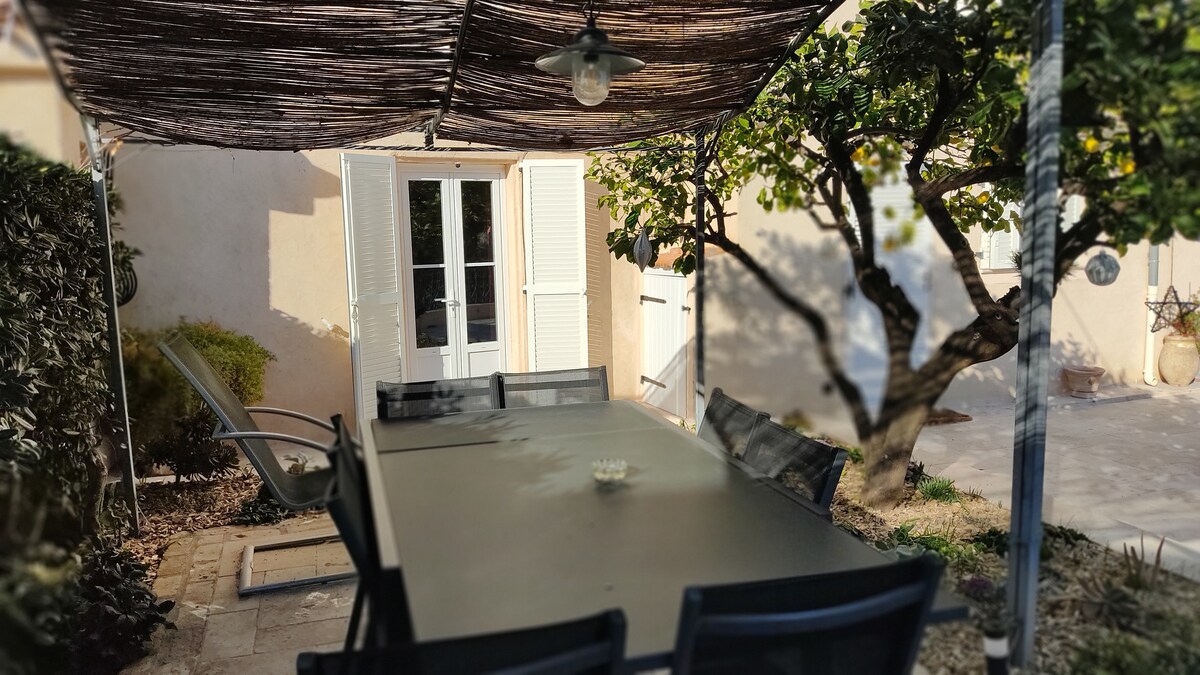 A shaded outdoor dining area is presented, featuring a large table surrounded by several chairs. A backdrop of light-colored walls and bright greenery creates a calm atmosphere. Sunlight filters through the canopy, enhancing the inviting space for relaxation or meals.