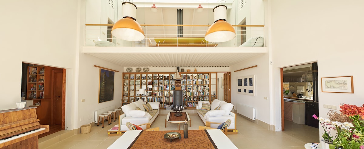 The spacious living area features high ceilings and large windows, allowing natural light to fill the space. Two comfortable sofas are positioned around a central coffee table, with a wood-burning stove adding warmth. A collection of framed photos adorns the wall, and a piano is visible nearby.