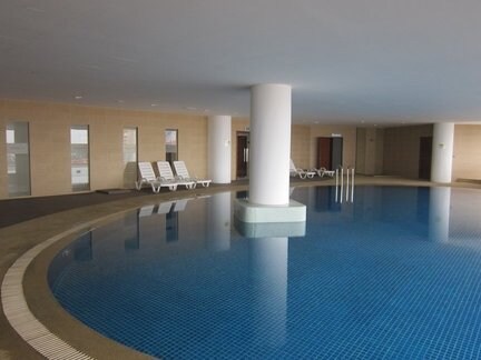 An indoor swimming pool area features clear blue water reflecting soft light. Lounge chairs are arranged along the sides, with tall columns providing structural support. Large windows allow natural light to fill the space, creating a serene environment for relaxation.