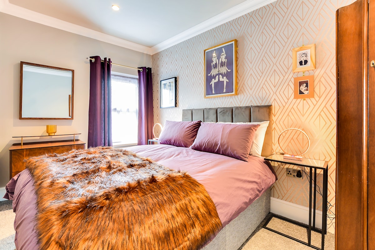 A well-appointed bedroom features a spacious bed adorned with purple bedding and a stylish fur throw. Gold accents are present in the wall art and decorative items. Soft lighting enhances the ambiance, while dark curtains frame the window, contributing to the room's cozy atmosphere.