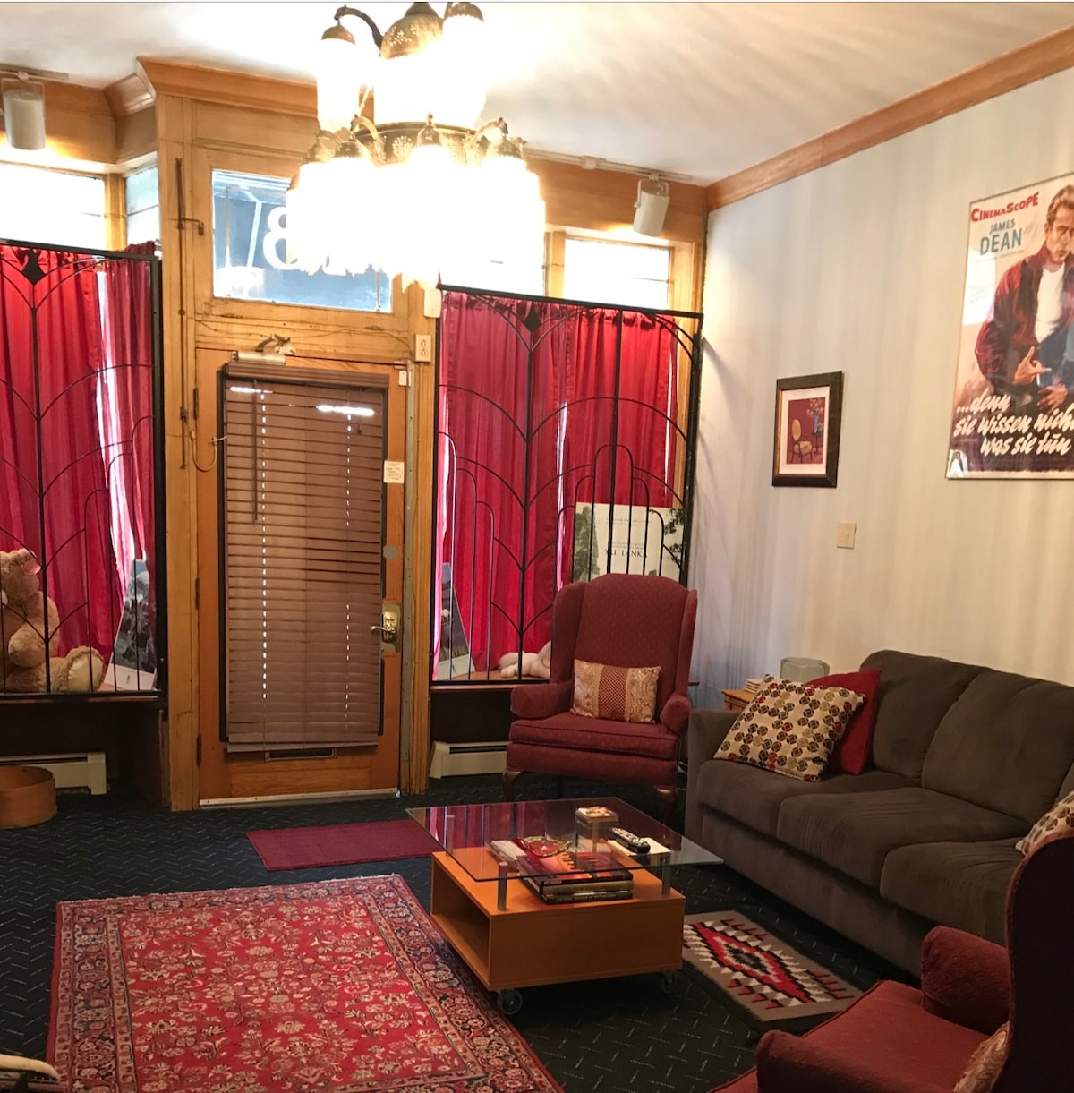 A spacious living area features large windows adorned with deep red curtains, allowing natural light to filter in. Comfortable seating options include two armchairs and a plush sofa, all set around a coffee table. A colorful area rug adds warmth to the space.
