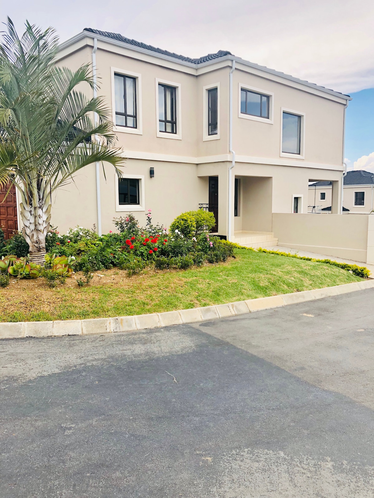 A well-maintained two-story house is presented, featuring a clean and modern façade. Lush greenery and colorful flowers enhance the front garden area. A smooth driveway leads to the entrance, providing a welcoming approach to the home.
