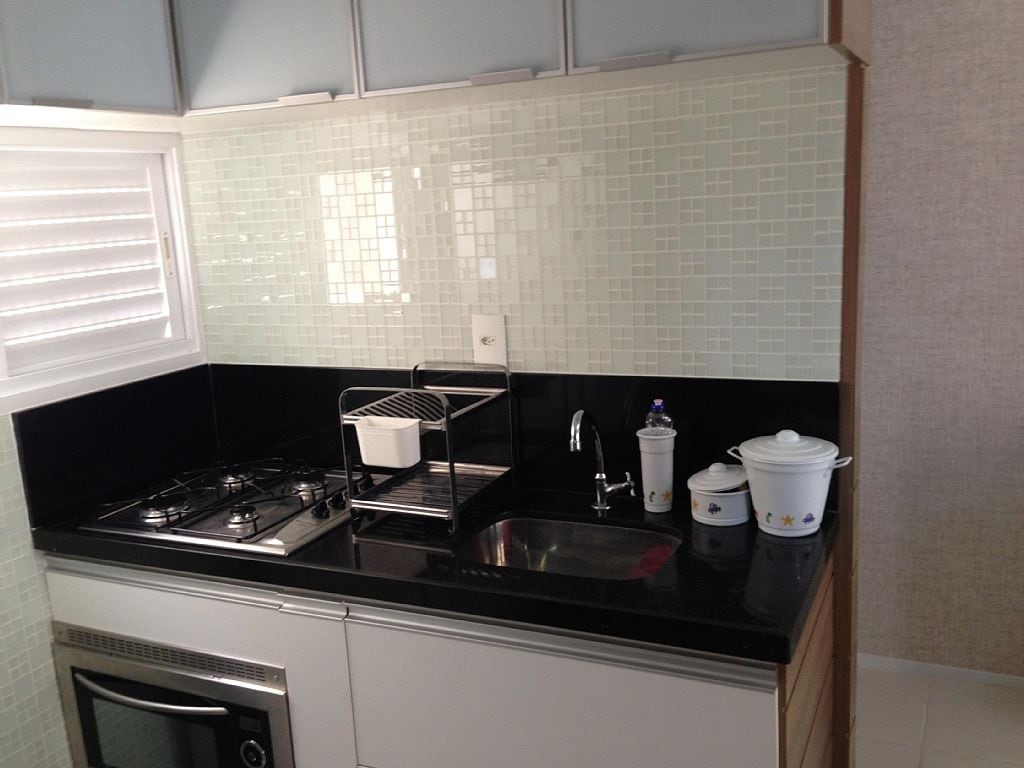A modern kitchen is presented with sleek cabinetry and a black countertop. Essential appliances, including a gas stove and sink, are visible. Storage containers and kitchen utensils are neatly arranged, with natural light entering through a window fitted with white shutters.