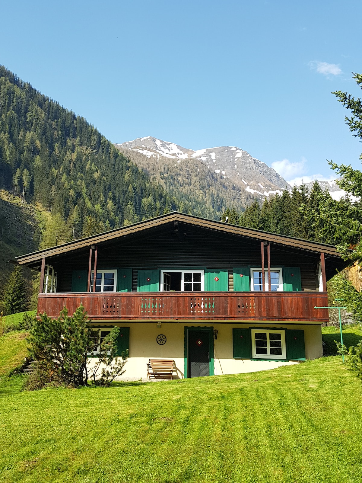 The two-story house features a rustic wooden façade with green shutters and a spacious balcony overlooking a grassy area. Surrounding mountains and trees provide a natural backdrop, enhancing the serene environment.