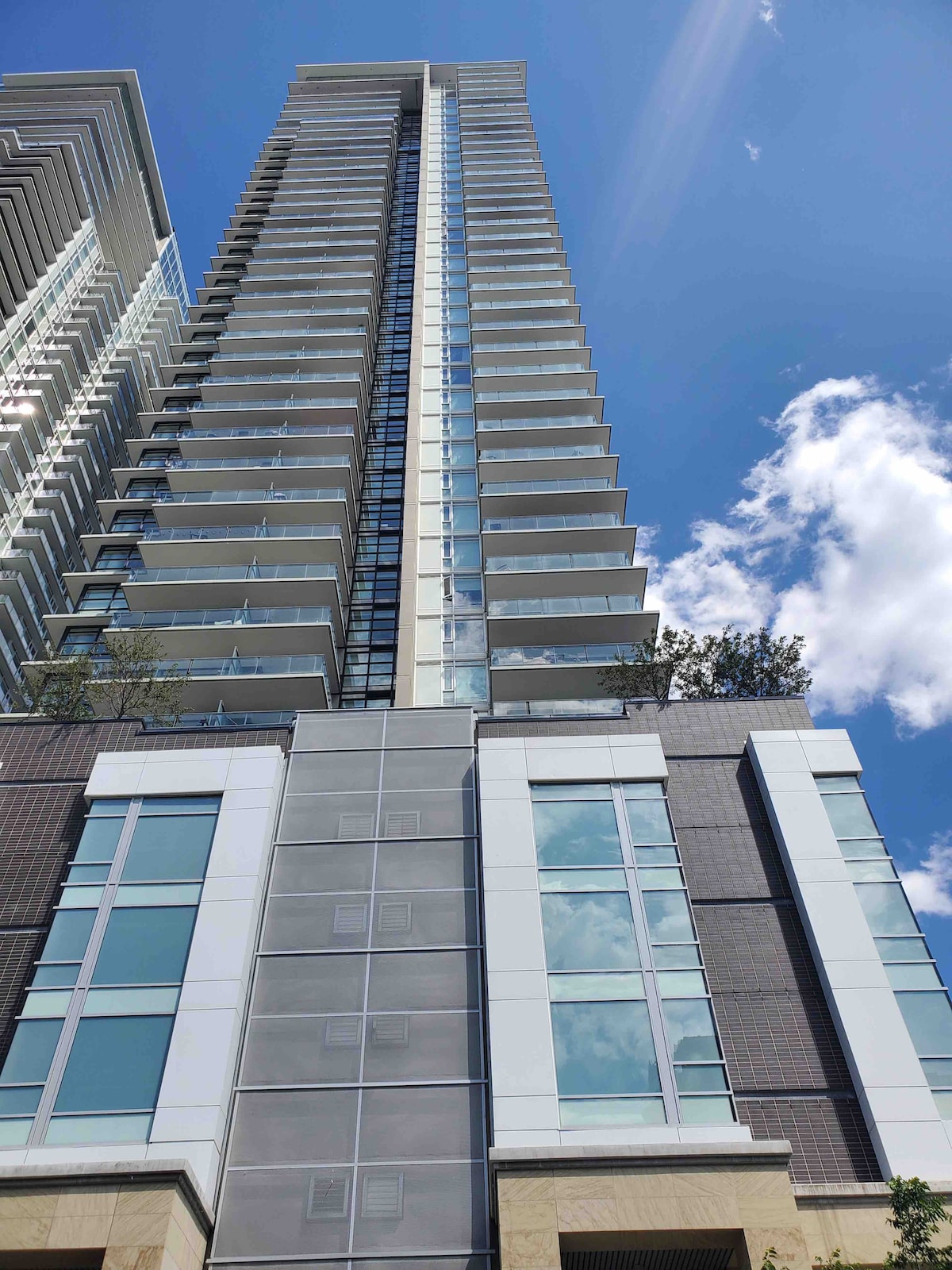 The image showcases a modern, tall residential building with multiple floors. Large glass balconies line the exterior, reflecting the sky. The structure's design features a combination of glass, stone, and metal elements, standing prominently against a backdrop of scattered clouds.