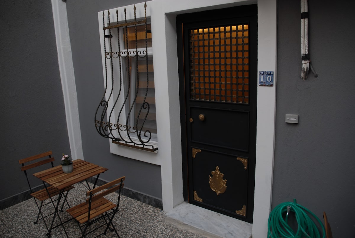 A small patio features a dark door with decorative brass accents and a window grate, providing an elegant entrance. Two wooden chairs and a compact table are arranged on the pebbled surface, accompanied by a potted plant, enhancing the welcoming outdoor space.