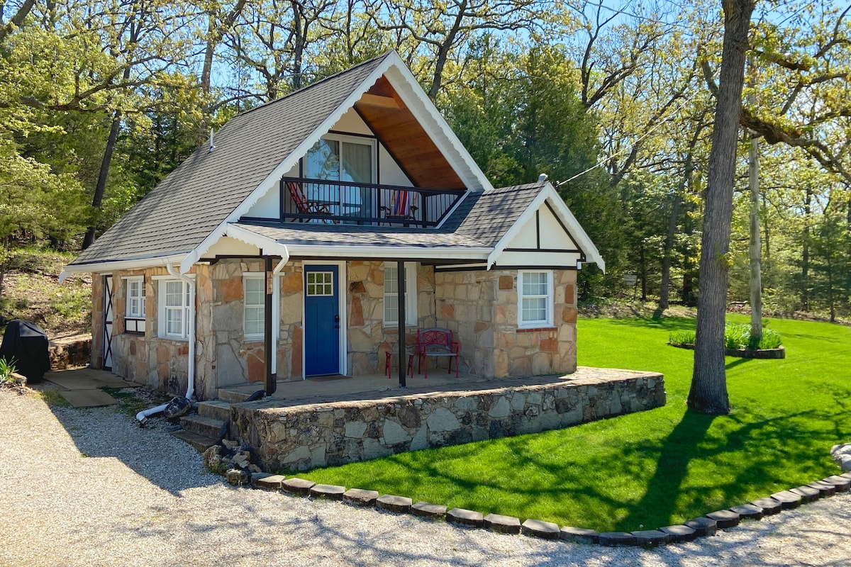 A charming cabin exterior is presented, featuring a stone facade and a pitched roof. A balcony is visible above the entrance, accompanied by a red chair set on the porch. Lush green lawn surrounds the cabin, with trees providing shade in the background.