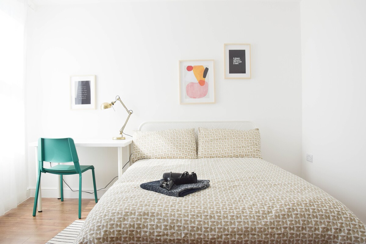 A cozy bedroom is highlighted by a spacious bed adorned with a geometric-patterned coverlet. A minimalist desk and chair are positioned nearby, accompanied by framed artwork on the walls. Natural light filters in through a window, enhancing the room's light and airy ambiance.