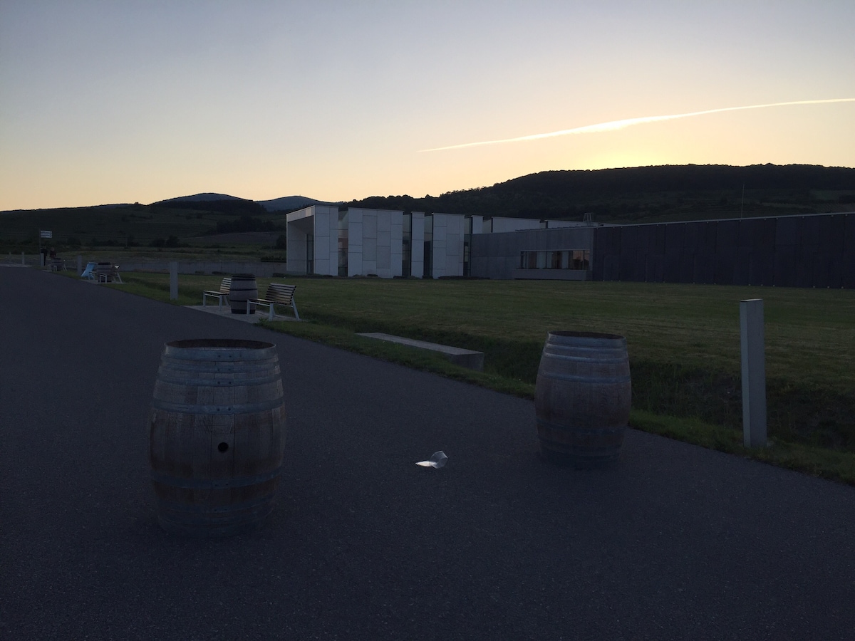 Two wooden wine barrels are positioned on a paved pathway, with a building in the background. The landscape features rolling hills under a dusky sky, illuminated by the fading light of sunset. Benches can be seen in the distance, surrounded by green grass.
