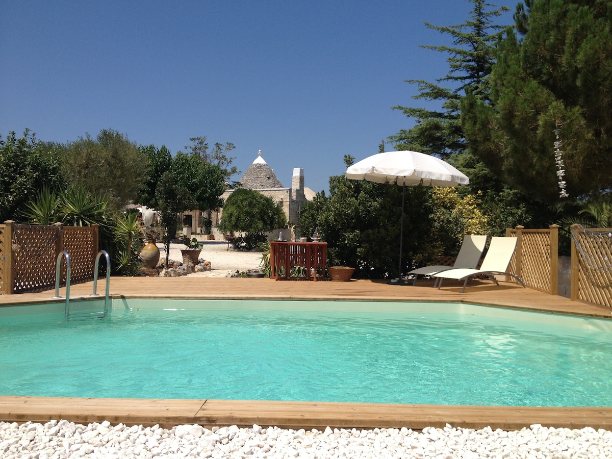 A swimming pool with clear water is surrounded by a wooden deck, featuring two lounge chairs and a large umbrella for shade. Lush greenery and shrubs provide a natural backdrop, with the trullo's distinctive conical roof visible in the distance.