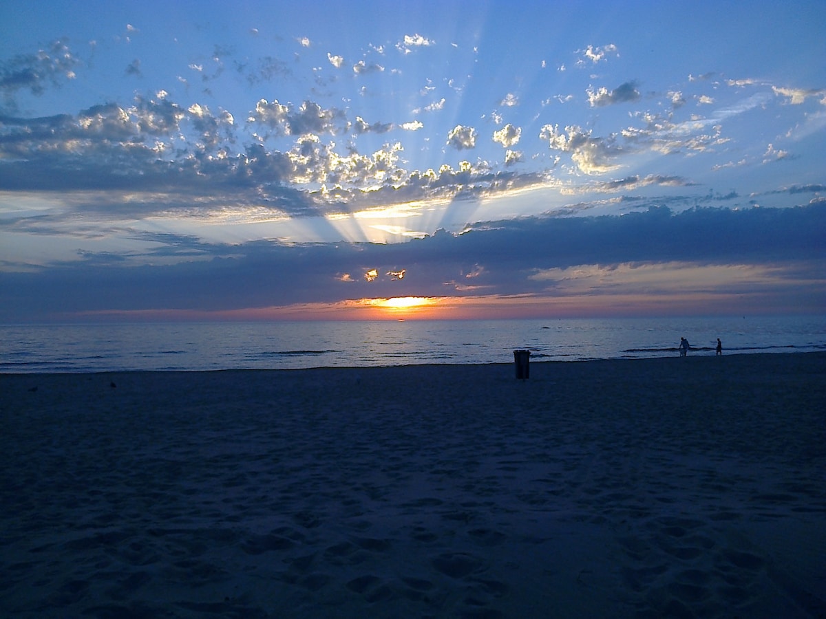 The image captures a serene beach scene during sunset, where the sun sinks below the horizon. Rays of light stretch across the cloudy sky, reflecting beautifully on the calm water. Silhouettes of two figures can be seen walking along the sandy shore.