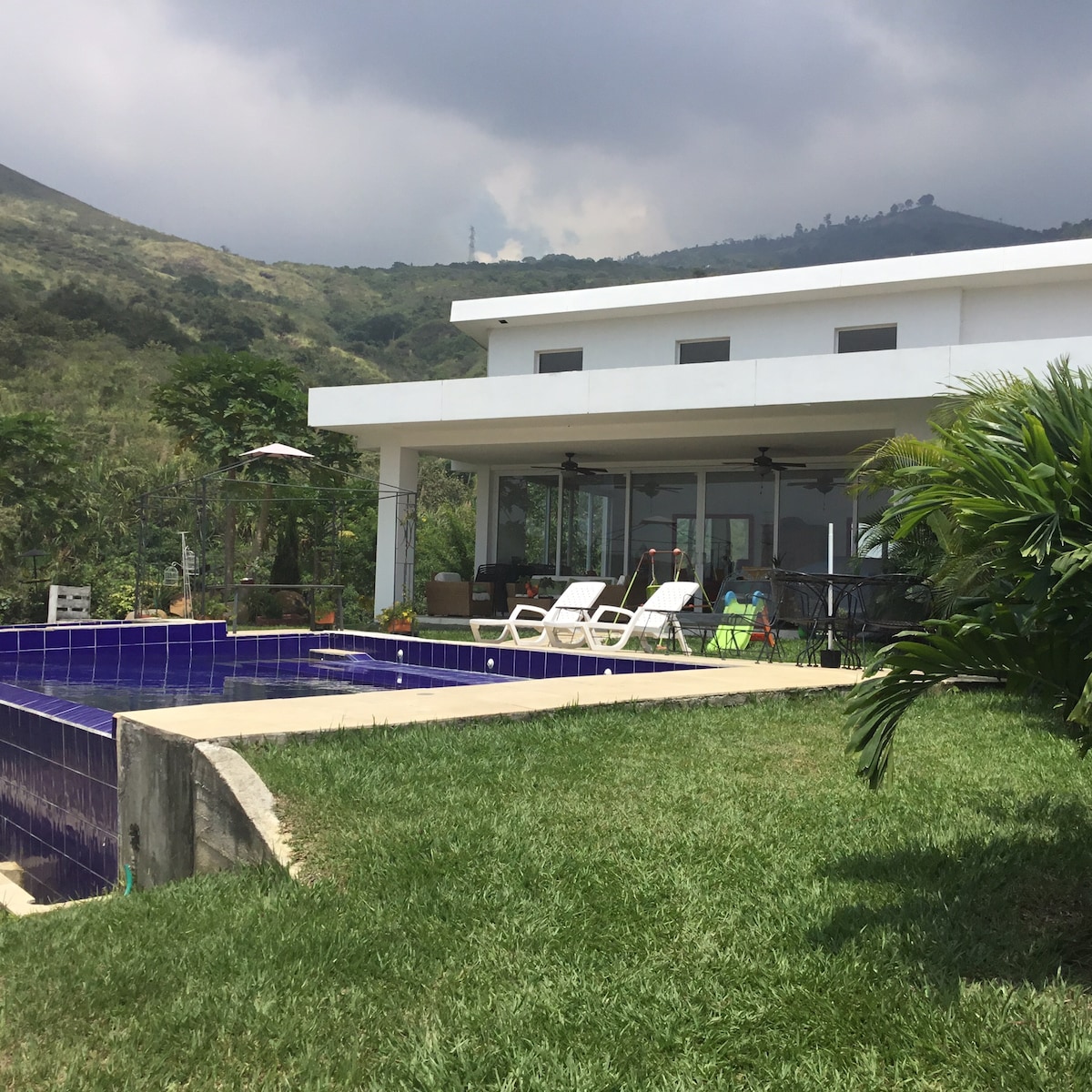 A modern home is surrounded by greenery, featuring a spacious outdoor area with a private pool. Several lounge chairs are positioned neatly by the poolside, with the backdrop of distant mountains and a cloudy sky, providing a serene atmosphere.