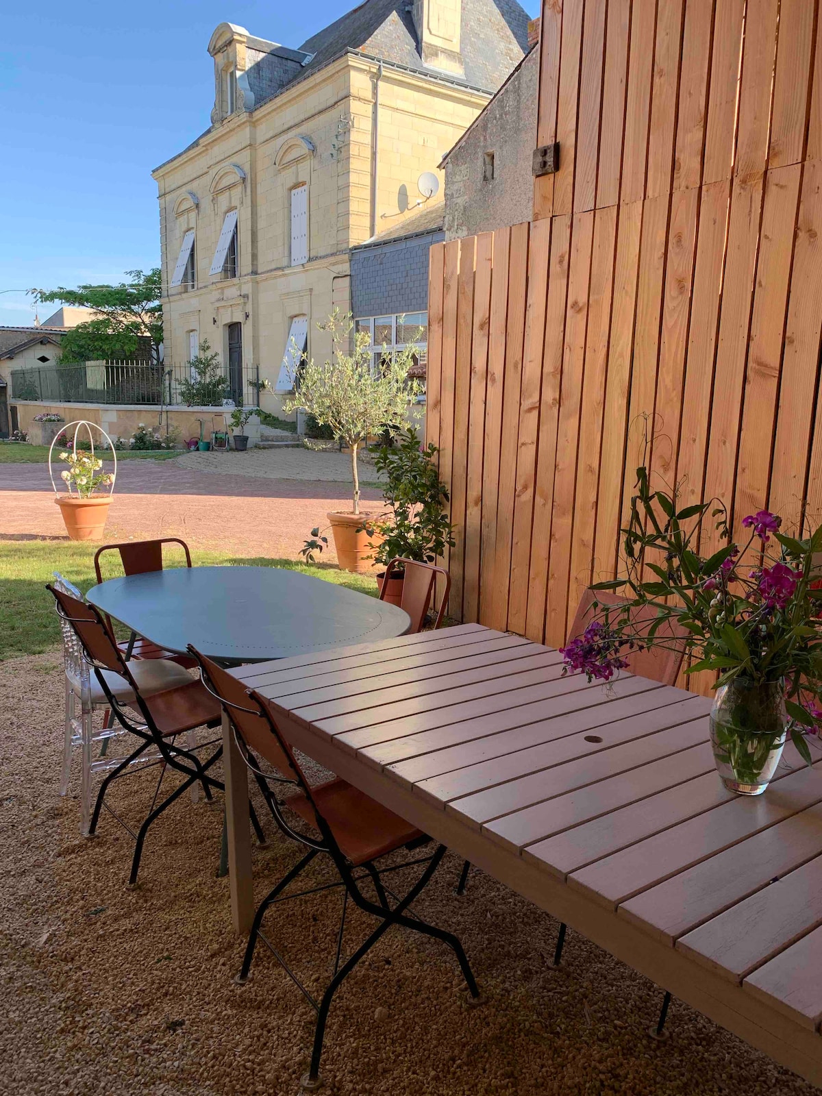 An outdoor terrace area is shown, furnished with a round glass table and a long wooden dining table surrounded by several chairs. Fresh-cut flowers are displayed in a vase, adding a touch of color. The background features a stone building, and greenery is visible.