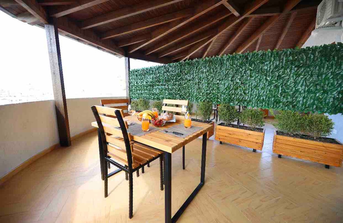A balcony area features a wooden dining table accompanied by two chairs. Potted plants line the space, providing a touch of greenery. The ceiling showcases a wooden structure, while a privacy screen made of artificial foliage enhances the tranquil setting.