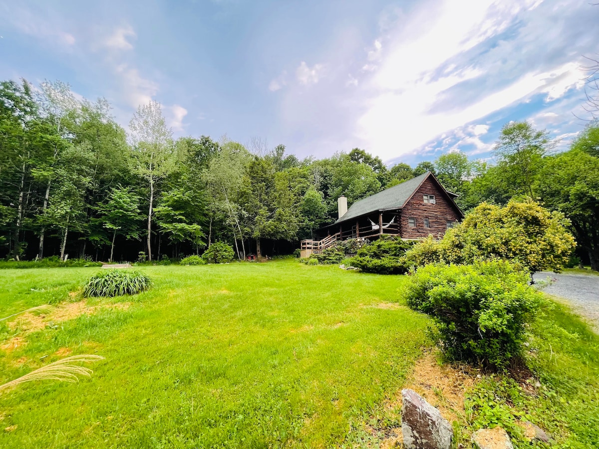 A log cabin is nestled among a lush green landscape, surrounded by trees. The structure features a sloped roof and a front porch with wooden railings. A grassy area leads to a gravel pathway for accessible entry.