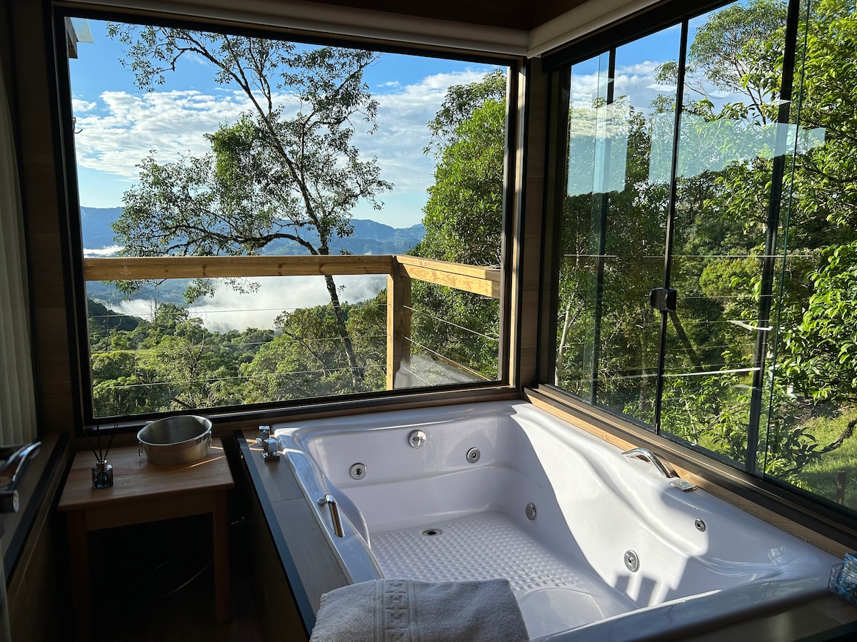 An inviting hydromassage bathtub is positioned by large windows, providing an expansive view of the surrounding mountains and lush greenery. Natural light filters through, enhancing the serene atmosphere. A small wooden table with a bowl is situated nearby.