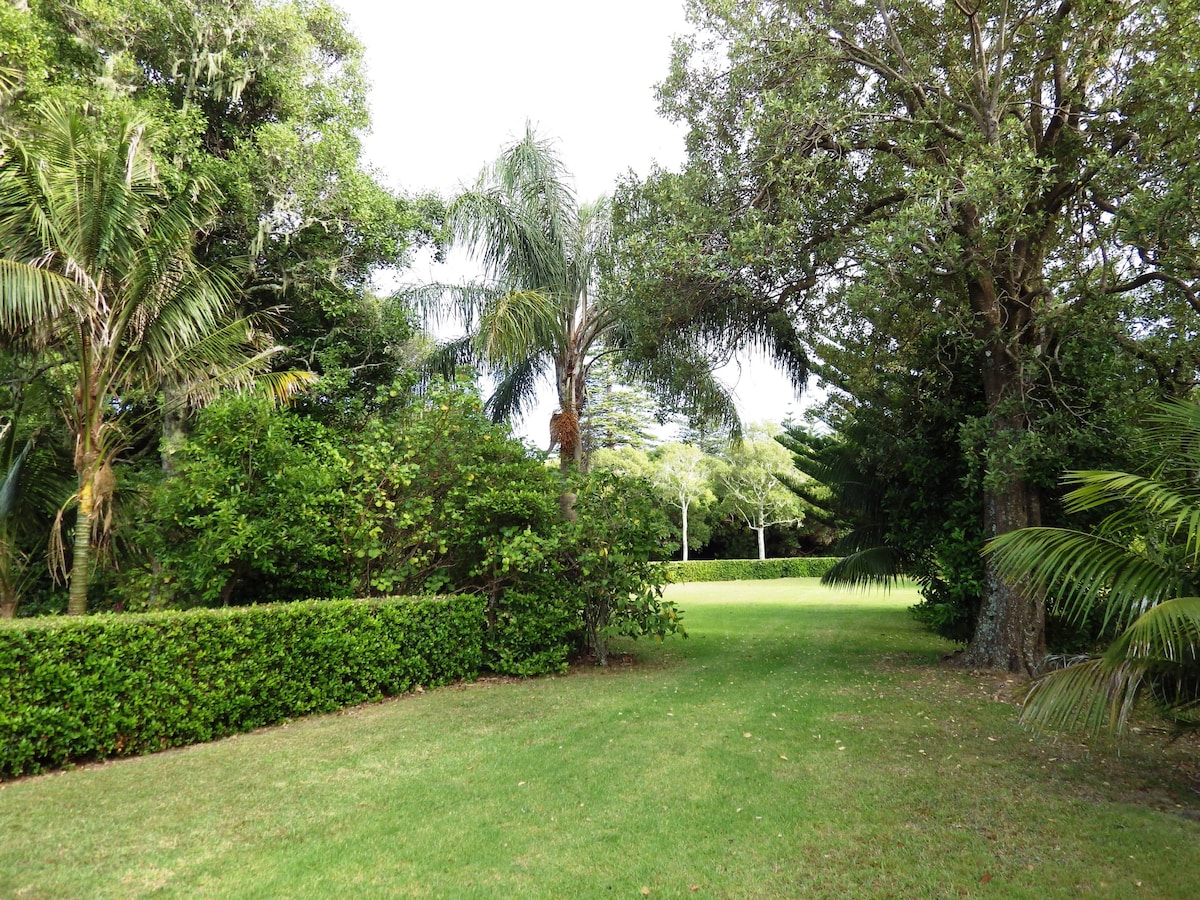 A lush, green expanse is surrounded by a variety of trees and shrubs, creating a serene garden setting. Palm trees and dense foliage are visible, adding to the natural aesthetics. A well-maintained lawn stretches into the distance under a partly cloudy sky.
