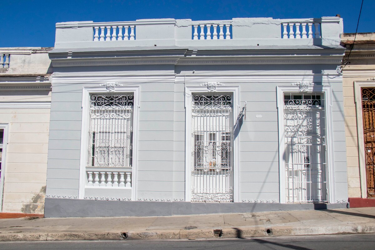 A colonial-style house is presented with a light gray façade, featuring intricate white wrought-iron detailing on the windows and doors. The structure includes a decorative cornice along the roofline, and the well-maintained sidewalk highlights the building's inviting entrance.
