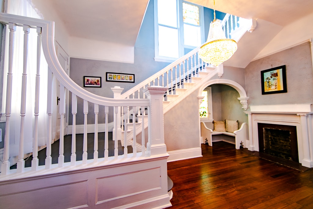 A grand staircase is featured with elegant railing, leading to an upper level. Natural light filters through large windows, illuminating the space. A cozy nook is visible beneath the staircase, along with a decorative fireplace, creating a welcoming entryway.