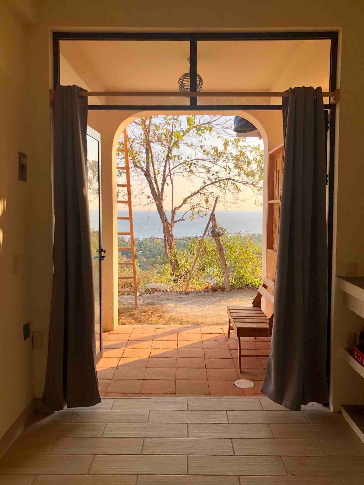 An open doorway is framed by two long curtains, revealing a scenic view of nature with a subtle gradient of colors in the sky. A small wooden bench is positioned just outside the door. The warm sunlight accents the earthy tones of the surrounding landscape.