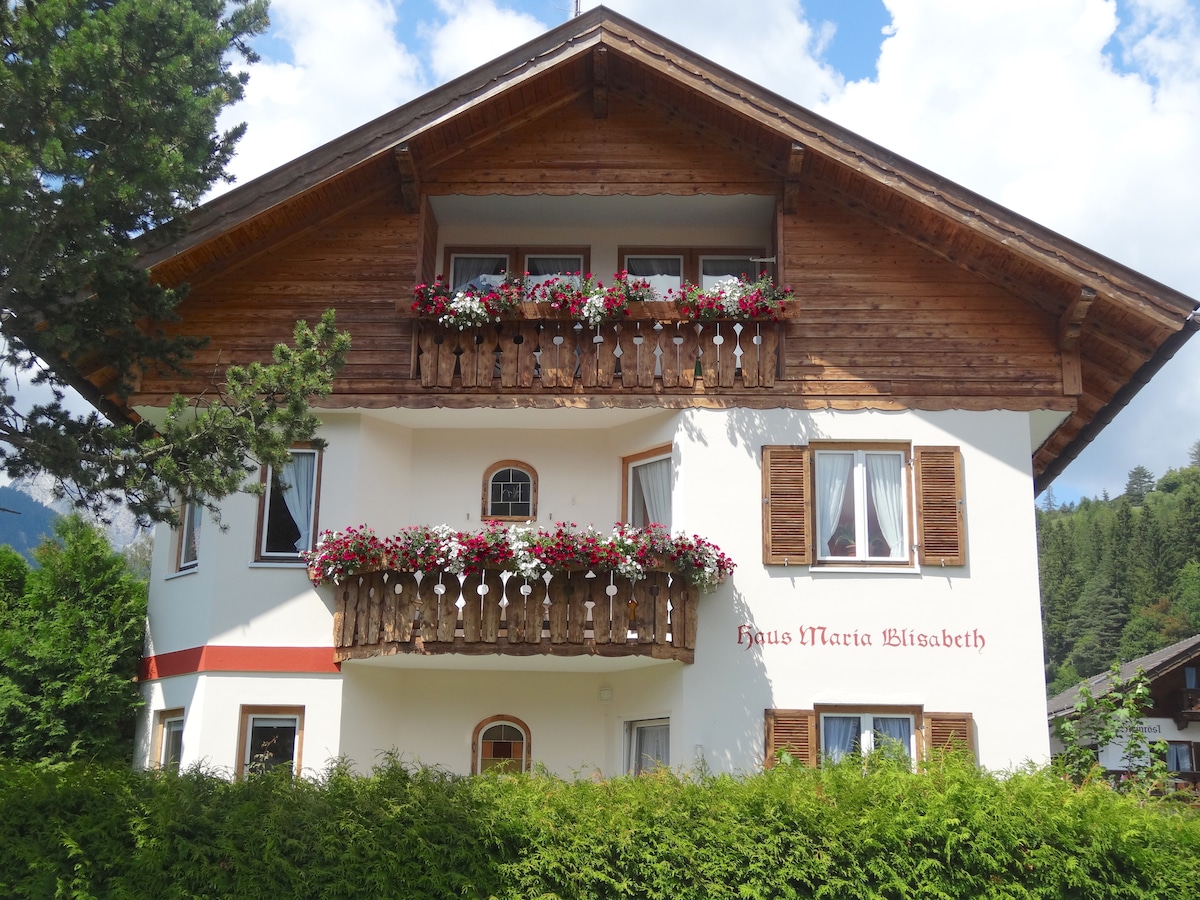 The charming guesthouse features a traditional alpine-style architecture with wooden balconies adorned with vibrant flower boxes. Large windows allow natural light to flood the interior, while greenery at the base enhances the welcoming appearance of the property.
