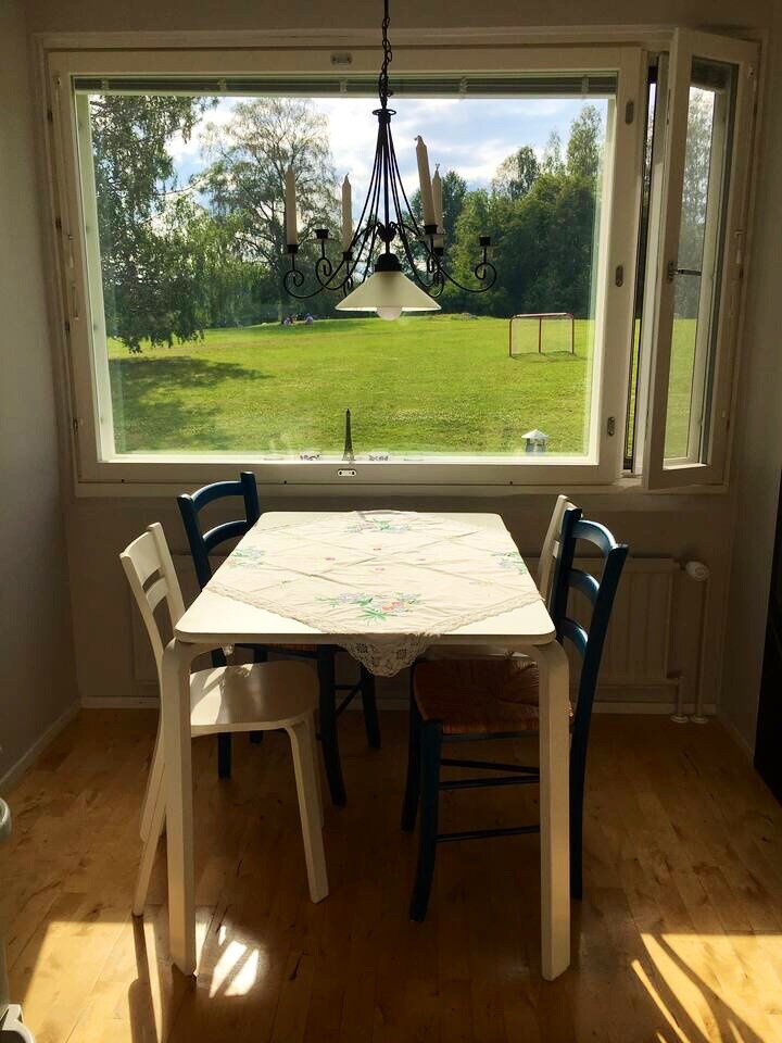 A dining area is positioned beside a large window, offering views of the green landscape outside. A table is set with a floral tablecloth, surrounded by two chairs – one white and one blue. Natural light illuminates the space, enhancing the calm atmosphere.