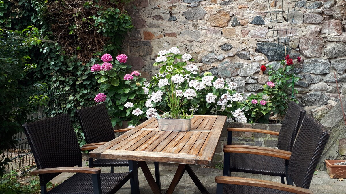 An outdoor dining area features a wooden table surrounded by six dark wicker chairs. Vibrant flowers in shades of pink and white bloom beside the stone wall, enhancing the tranquil setting. Lush greenery provides a natural backdrop to this inviting space.
