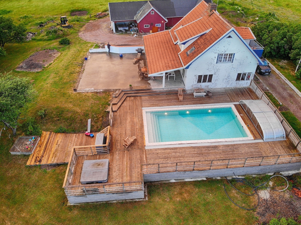 An aerial view presents the property featuring a large wooden deck surrounding a swimming pool and jacuzzi. The house, with a red exterior and orange tiled roof, is complemented by grassy surroundings. Space for outdoor seating is visible, along with a barbecue area.