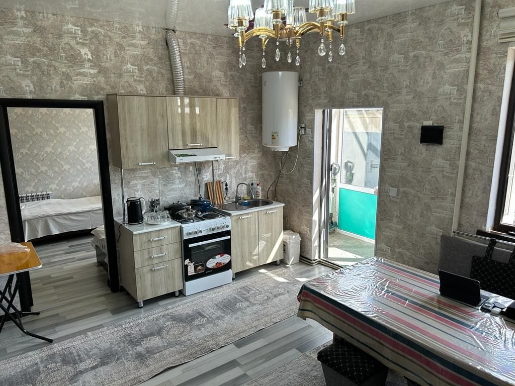 The spacious kitchen area features modern cabinetry and appliances, including a stove and a sink. A dining table with a striped tablecloth is visible, and a chandelier hangs from the ceiling. Light enters through nearby windows, illuminating the area.