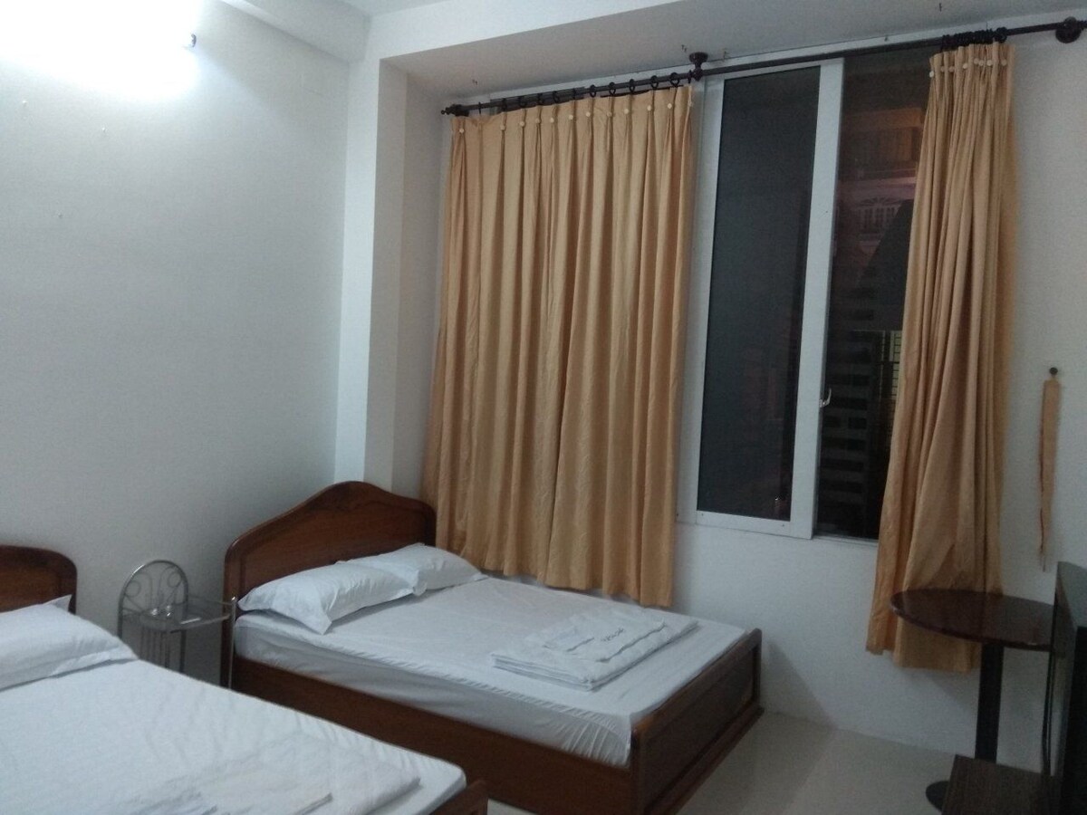 A bedroom is seen featuring two wooden beds, each with neatly arranged white linens. A large window is draped with light beige curtains, allowing natural light to fill the space. A small table and television are present, enhancing the functional layout.