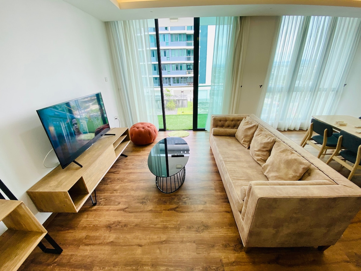 The living room features a spacious couch, a flat-screen TV positioned on a light wooden media console, and a round coffee table. Floor-to-ceiling windows provide abundant natural light and reveal the view of a green outdoor area. A cozy seating option is available nearby.