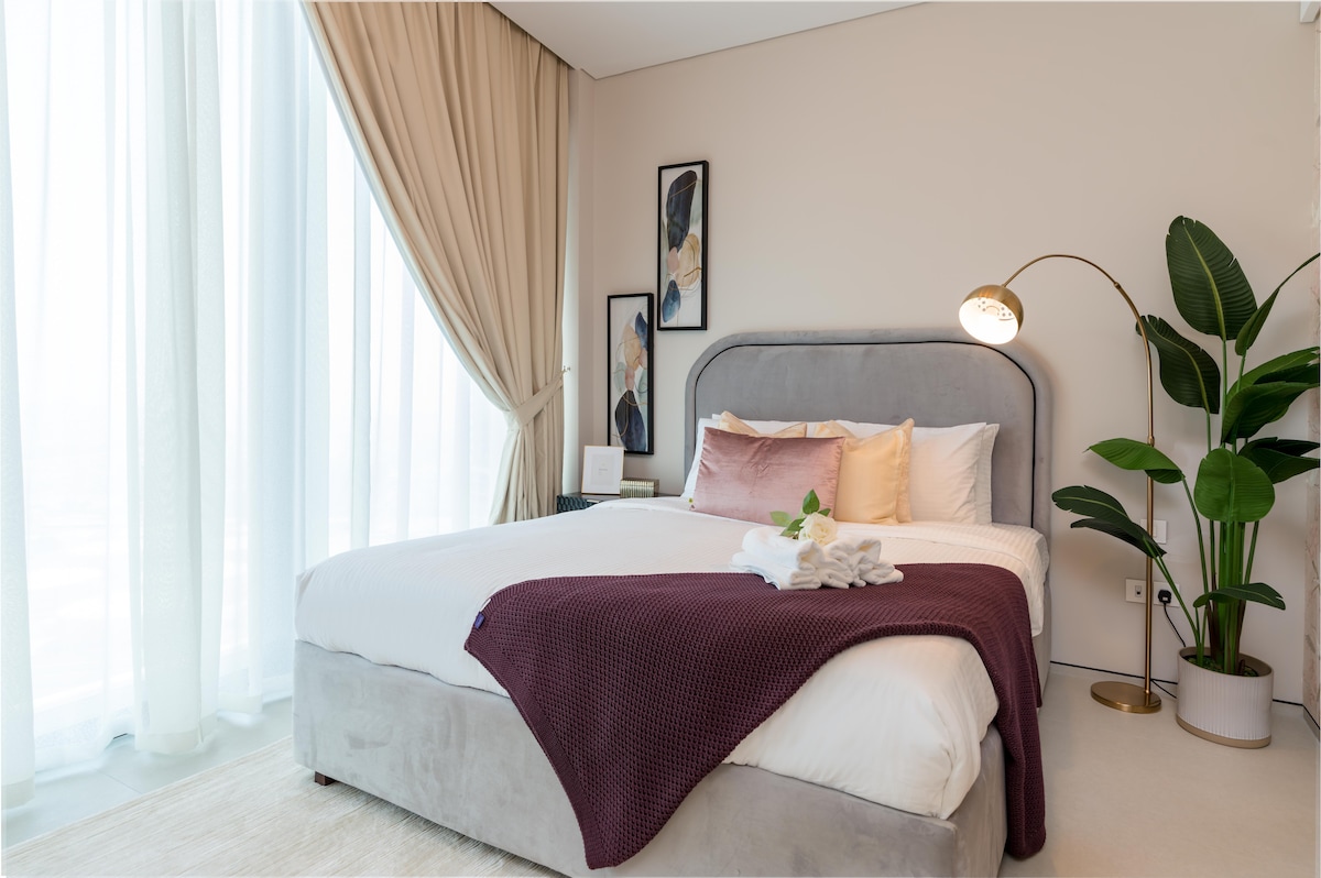 A spacious bedroom features a plush bed adorned with decorative pillows and a cozy throw. Floor-to-ceiling windows draped in sheer curtains allow natural light to fill the room, while a modern lamp and a potted plant add a touch of elegance to the space.
