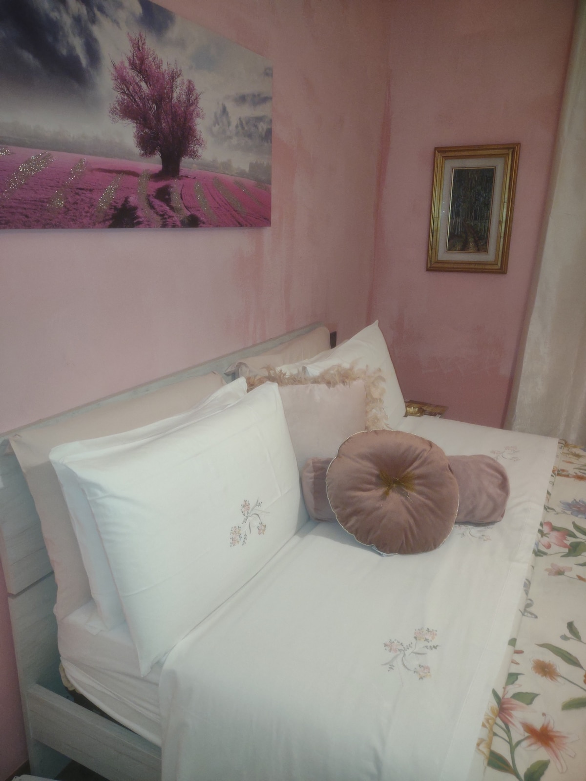 A spacious bed is adorned with embroidered white bedding and decorative pillows in soft hues. A floral-patterned throw adds a touch of color. Above the bed, a serene landscape photograph captures a flowering tree, complementing the room's pink walls and elegant decor.