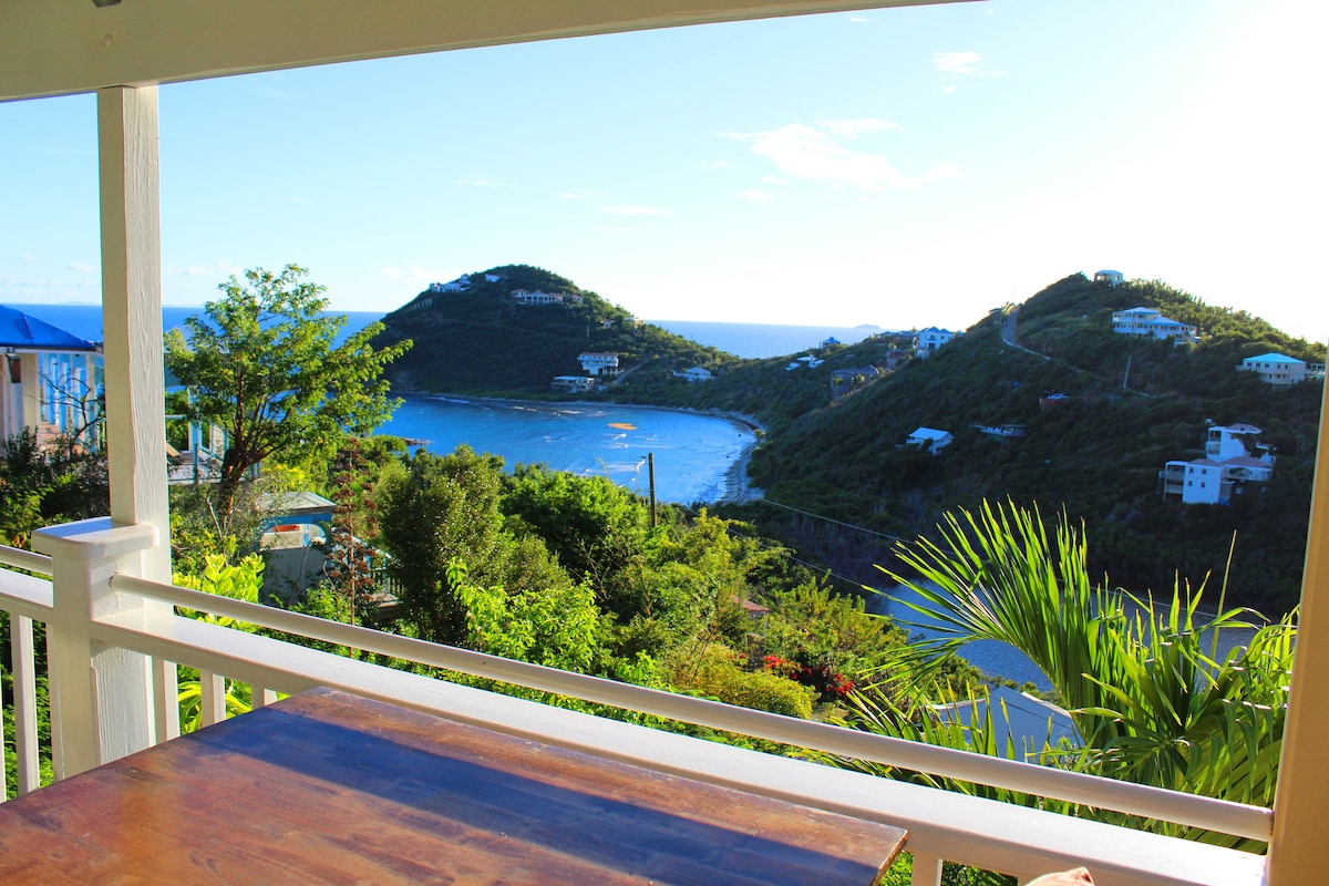 A peaceful view is captured from the covered deck, showcasing lush green hills descending towards turquoise waters. The horizon features distant hills and a clear sky, enhancing the natural beauty of the surrounding landscape.