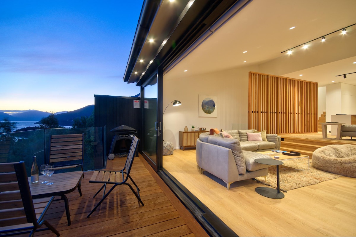 An outdoor deck area features two folding chairs and a small table, inviting relaxation with scenic views in the background. The interior space showcases comfortable seating, warm wood accents, and soft lighting, creating a cozy ambiance. Lake Wakatipu and mountains are visible beyond the large glass doors.