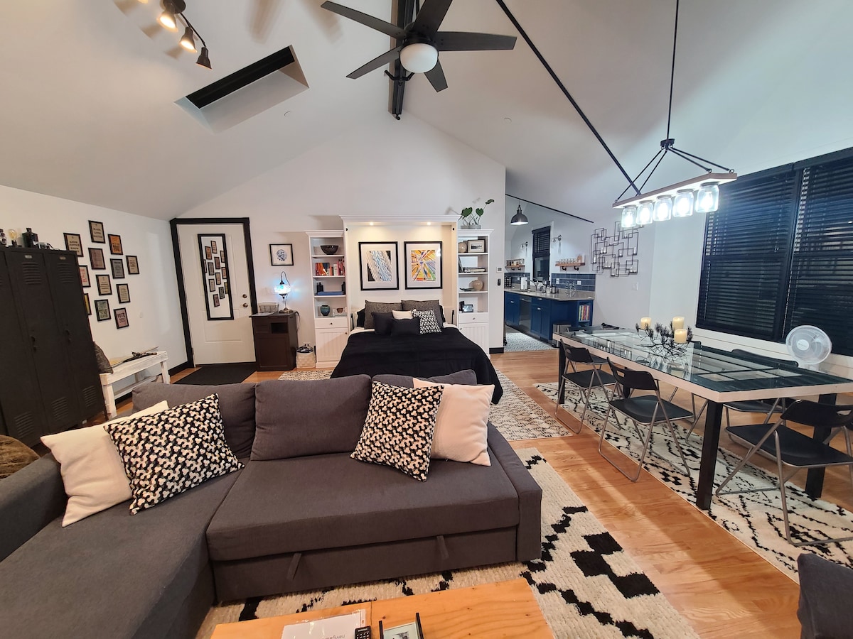 An open living area is highlighted by a spacious dark sofa adorned with decorative cushions, complemented by a wood coffee table. A large, comfortable bed occupies one side, while a dining area with a glass table and chairs is visible. Art pieces are displayed on the walls.