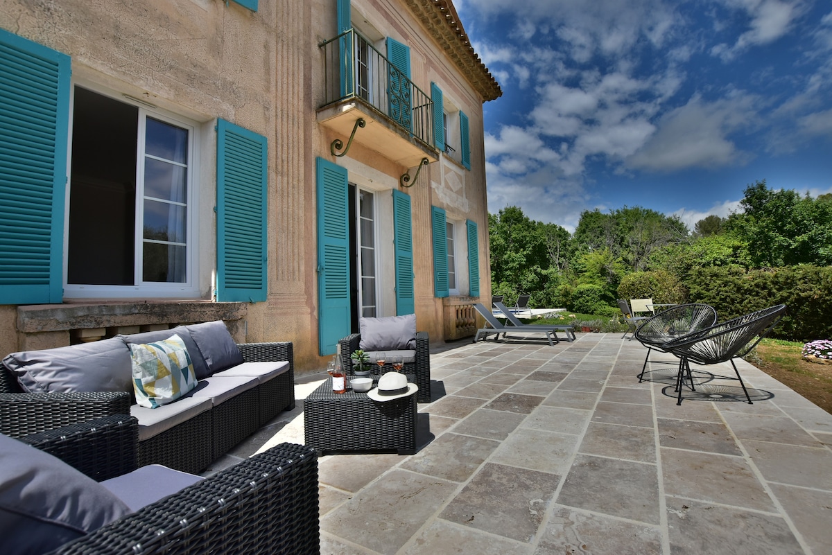 A spacious outdoor terrace features comfortable seating arrangements, including wicker furniture with soft cushions. Bright blue shutters frame large windows, while a well-maintained garden is visible in the background. The area is bathed in natural light under a partly cloudy sky.