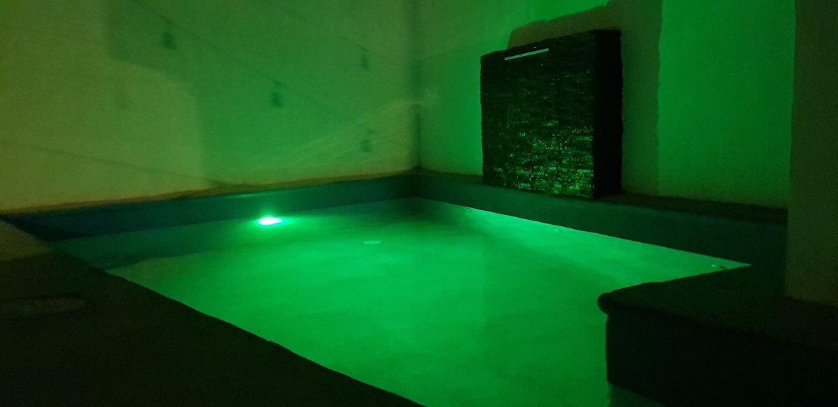 A serene pool area is illuminated with soft green lighting, creating a calming atmosphere. A stone waterfall feature adds texture to the space, while smooth surfaces and gentle curves define the pool's design. The overall ambiance suggests a relaxing environment for leisure.