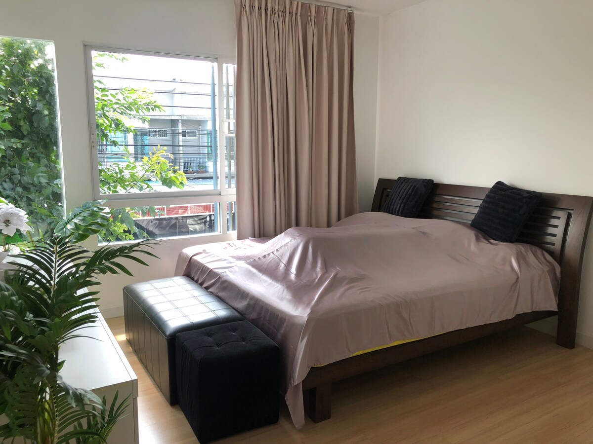 A bedroom features a spacious bed covered with a light-colored duvet. A dark bench is positioned at the foot of the bed. Natural light enters through large windows framed by sheer curtains, while a small plant adds a touch of greenery to the room.