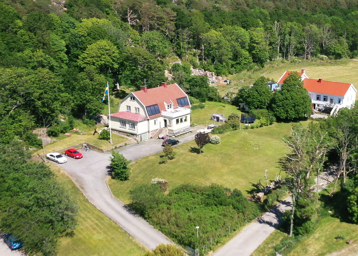 An elevated view reveals a charming property surrounded by greenery, featuring a large lawn and various trees. Two buildings are visible, one with a red roof and solar panels, the other a white structure, with several vehicles parked along a winding driveway.