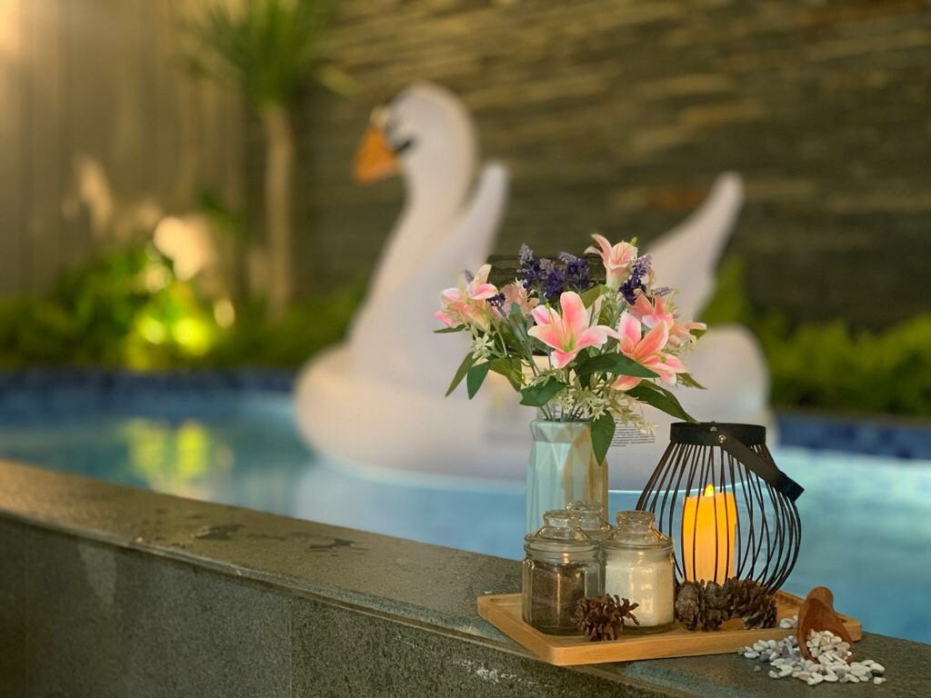 A tranquil pool area is complemented by a decorative tray featuring jars, a candle, and natural accents. In the background, a large inflatable swan floats gently on the water surrounded by lush greenery.