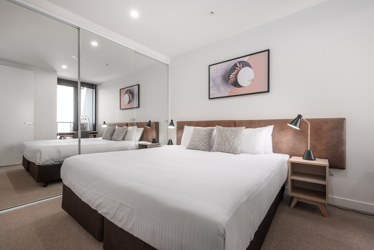 A bright bedroom features a spacious bed with crisp white linens and decorative pillows. A large mirror reflects the room, enhancing the sense of space. A bedside table with a lamp is positioned on each side of the bed. Artwork adorns the wall above the headboard.