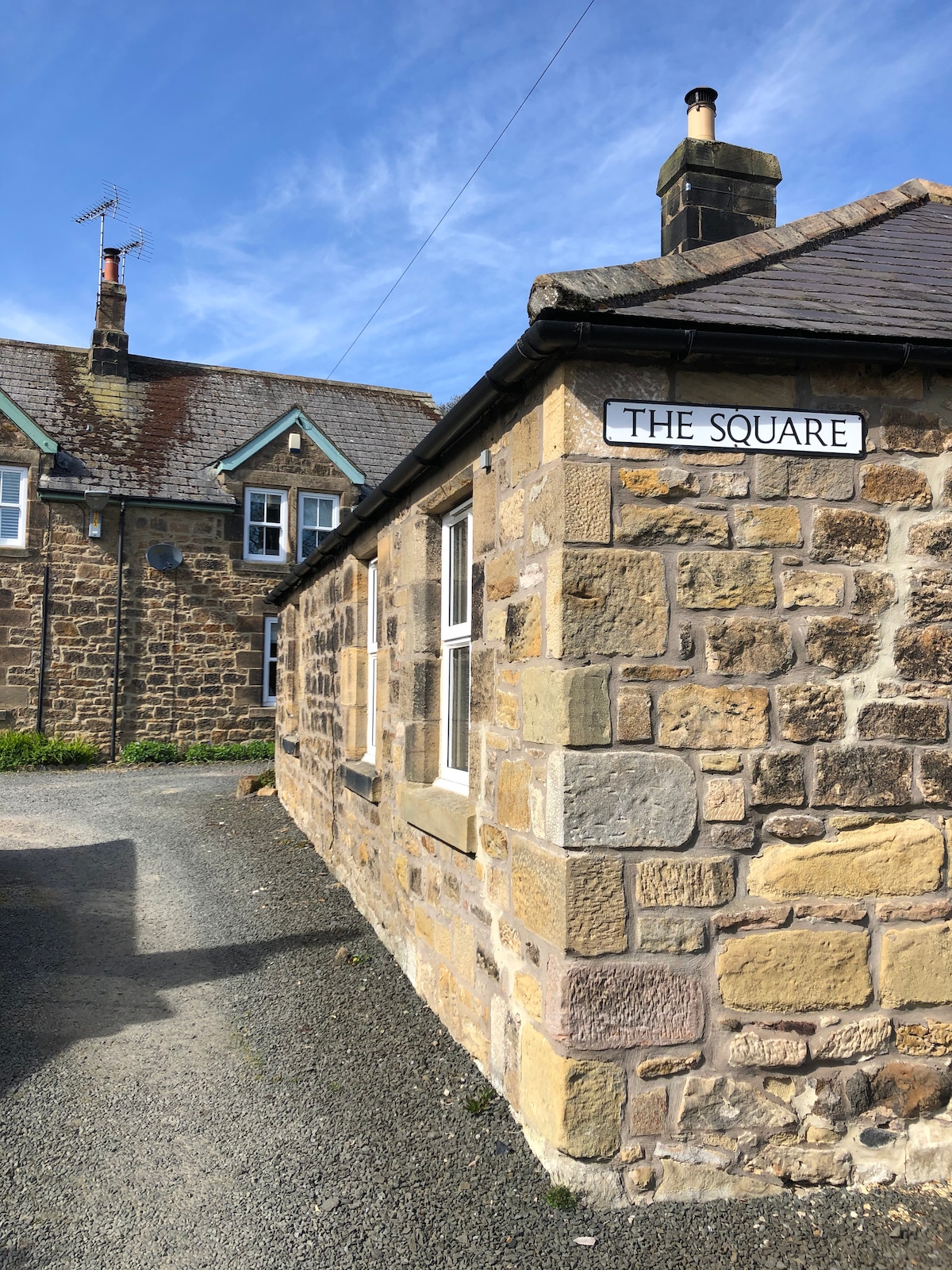 A charming stone cottage features a sign reading 'The Square.' The textured stone exterior showcases a blend of colors, while a clear blue sky is visible above. The gravel path leading to the cottage is framed by neatly arranged buildings in the background.